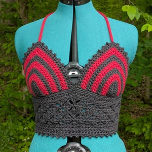 May include: A handmade crochet top in a halter style, displayed on a mannequin. The top features a striking design with alternating red and dark gray chevron stripes on the bust and a detailed dark gray band at the waist. The halter straps are red.