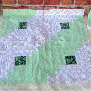 May include: A green and white quilt block with a log cabin pattern. The block is made up of squares of different sizes, with the smallest square in the center. The squares are arranged in a spiral pattern, with the largest square on the outside.