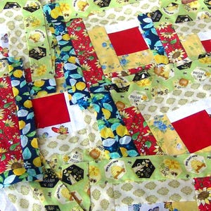 May include: A patchwork quilt with a repeating pattern of red squares surrounded by yellow, green, and blue fabric. The fabric features various patterns, including lemons, bees, and sunflowers.