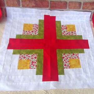 May include: A white fabric square with a red, green, and yellow patchwork design in the center. The design is a series of squares within squares, creating a log cabin pattern.