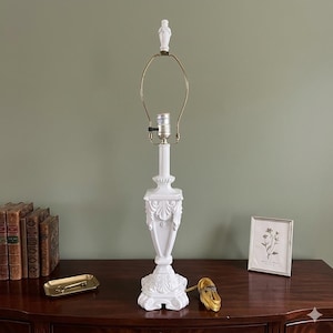 Può includere: Una lampada da tavolo bianca alta con base quadrata decorata con intagli a foglia, una colonna centrale sottile e un'arpa in metallo color ottone. La lampada include un terminale bianco sulla sommità e un cavo elettrico color oro.