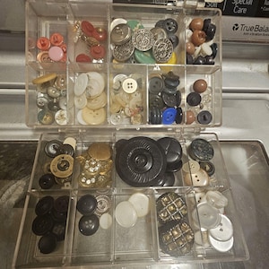 May include: A clear plastic organizer box filled with a variety of sewing buttons in assorted shapes, sizes, and colors including black, white, gold, red, and blue. The buttons are sorted into individual compartments within the two-tier container.