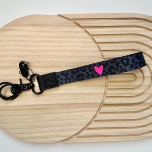 Könnte beinhalten: Ein Schlüsselanhänger aus Stoff mit schwarz-grauem Leopardenmuster und einem kleinen, leuchtend pinken Herzsymbol. Das Band ist an einem schwarzen Metallverschluss und einem Schlüsselring befestigt, mit einem schwarzen Kunststoffverbinder.