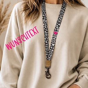 Könnte beinhalten: Ein Stoff-Lanyard mit einem schwarz-braun-weißen Leopardenmuster und einem kleinen pinken Herz-Symbol. Das Lanyard verfügt über ein beiges Kunstleder-Verbindungsstück und einen drehbaren Karabinerhaken aus dunklem Metall. Der Text WUNSCHTEXT ist sichtbar.