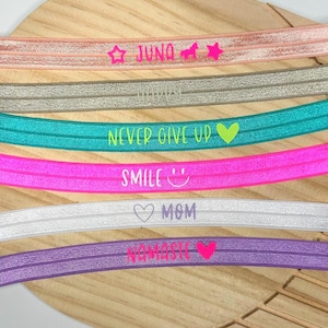 Könnte beinhalten: Sechs elastische Stirnbänder in verschiedenen Farben wie Pfirsich, Gold, Türkis, Pink, Weiß und Lila. Jedes Band ist mit Text und Symbolen bedruckt: JUNA mit Stern und Einhorn, HAPPY, NEVER GIVE UP mit Herz, SMILE mit Gesicht, MOM mit Herz und NAMASTE mit Herz.