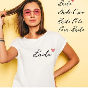 Peut inclure: Un t-shirt blanc avec le mot "Bride" en écriture noire et un petit cœur rose. L'image affiche également les phrases "Bride Crew", "Bride To Be" et "Team Bride". Le texte "JGA SHIRTS - DIY" est également visible.