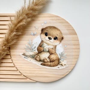 Könnte beinhalten: Eine runde Holzplatte mit einer gedruckten Illustration eines braunen Otters, der an einem sandigen Ufer sitzt und eine Muschel hält. Der Otter hat große dunkle Augen und ist von kleinen Seesternen, Blasen und hellgrünen Wasserpflanzen umgeben.