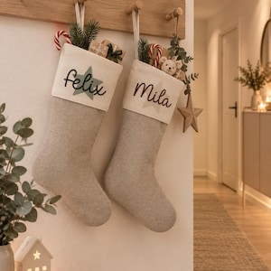 Könnte beinhalten: Zwei beigefarbene Weihnachtsstrümpfe aus Leinen hängen an einer hölzernen Wandleiste. Der linke Strumpf zeigt den Namen 'felix' in schwarzer Schreibschrift über einem grünen Stern. Der rechte Strumpf zeigt den Namen 'Mila' über einem beigen Stern.