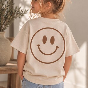 Könnte beinhalten: Ein beiges Kurzarm-T-Shirt mit einem großen braunen Smiley-Gesicht auf der Rückseite. Das T-Shirt hat einen Rundhalsausschnitt und eine lockere Passform, kombiniert mit einer hellblauen Jeans mit sichtbaren Gesäßtaschen und einem Bund.