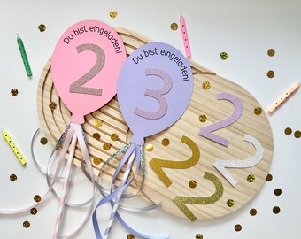 May include: Pink and purple balloon-shaped birthday decorations with glitter numbers 2 and 3. The balloons have ribbons and the text "Du bist eingeladen!". Additional glitter numbers and candles are scattered on a wooden surface.