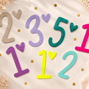 Könnte beinhalten: Eine Sammlung von bunten, glitzernden und matten Zahlen und Herzformen, die auf einer hellen Oberfläche angeordnet sind. Das Set enthält die Zahlen 2, 3, 5, 1, 1, 1 und 2 in verschiedenen Farbtönen wie Beige, Lila, Türkis, Pink, Gelb und Hellblau.