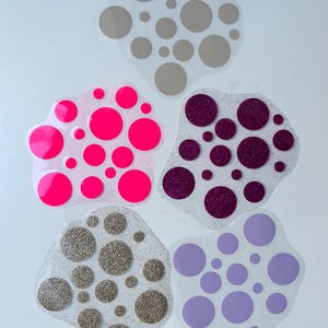 Könnte beinhalten: Fünf unregelmäßige, durchscheinende, klecksförmige Klebefolien mit verschiedenen kreisförmigen Mustern. Die Designs umfassen verschieden große Kreise in Neonpink, Helllila, Grau, glitzerndem Dunkellila und glitzerndem Gold.