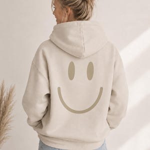 Könnte beinhalten: Ein beigefarbener Kapuzenpullover mit einem großen, mittig platzierten, hellbraunen Smiley-Gesicht auf der Rückseite. Der Hoodie hat lange Ärmel, eine Kapuze mit Kordelzug und eine lockere Passform, kombiniert mit einer hellblauen Jeans.