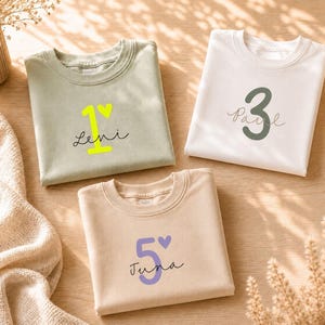 Könnte beinhalten: Drei gefaltete Rundhals-Sweatshirts in Salbeigrün, Weiß und Beige. Das salbeigrüne Shirt zeigt eine neongelbe Zahl 1, ein Herz und den Namen Leni. Das weiße Shirt hat eine dunkelgrüne Zahl 3 und den Namen Paul. Das beige Shirt zeigt eine violette Zahl 5, ein Herz und den Namen Juna.