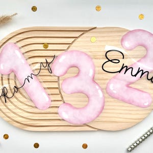 Könnte beinhalten: Ein ovales Holzbrett mit Rillen zeigt drei große, glänzende, hellrosa Zahlen: 1, 3 und 2. Der Name Romy ist in schwarzer Schreibschrift über der Zahl 1 und der Name Emma in schwarzer Schreibschrift über der Zahl 2 angebracht.