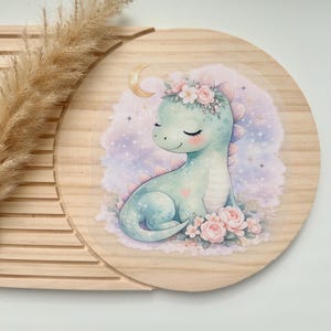 Könnte beinhalten: Runder Holzschild mit einer Aquarellillustration eines niedlichen, hellblauen Dinosauriers mit Blumenkrone, der inmitten von rosa Rosen sitzt. Ein Mond und Sterne sind im Hintergrund. Das Schild ist aus hellem Holz gefertigt.