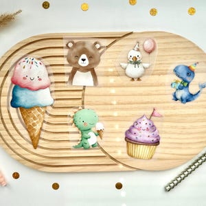 Könnte beinhalten: Ein Set aus sechs bunten Aufklebern mit einer lächelnden Eiswaffel, einem braunen Bären, einer weißen Ente mit Luftballon, einem blauen Dinosaurier, einem grünen Dinosaurier und einem lila Cupcake auf einem hellen, ovalen Holztablett mit Rillen.