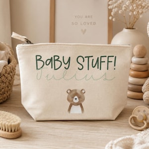 Op de afbeelding: Een crèmekleurig canvas tasje met ritssluiting voorzien van de donkergroene tekst "BABY STUFF!" boven de naam "Julius" in een lichtgroen cursief lettertype. Onder de tekst staat een kleine illustratie van een bruine beer.
