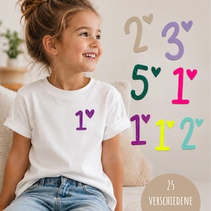 Könnte beinhalten: Ein weißes T-Shirt mit einer lila Glitzer-Eins und einem kleinen Herzsymbol. Neben dem T-Shirt sind verschiedene Zahlen in unterschiedlichen Farben und Schriftarten abgebildet, darunter Beige, Lila, Dunkelgrün, Pink, Gelb und Hellblau.