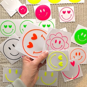 Puede incluir: Una colección de pegatinas de vinilo con varios diseños de caras sonrientes en colores neón y sólidos. Las pegatinas incluyen sonrisas clásicas, ojos en forma de corazón y contornos de flores en tonos rosa, naranja, verde, amarillo y negro.