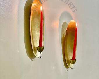 May include: Two wall-mounted candle holders with a gold finish. Each holder features a red candle, lit with a flame. The holders are mounted on a white wall, casting shadows. The text "MOROCCOALLARTS" is visible.