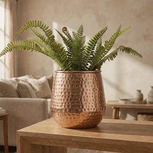 Puede incluir: Una maceta redonda de metal martillado color cobre con una abertura ancha y una base cónica. La maceta contiene una planta de helecho artificial verde y frondosa con múltiples frondas que se extienden desde el centro del recipiente.