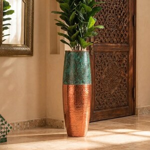 May include: A tall, cylindrical floor vase featuring a hammered metal texture. The upper half displays a weathered, oxidized teal patina, while the lower half showcases a polished, warm copper finish. A green leafy plant is placed inside the vase.