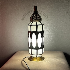 Op de afbeelding: Een sierlijke tafellamp met een messing voet en top, met een wit en zwart geometrisch ontwerp. De lamp heeft een cilindrisch lichaam met boogvormige uitsparingen en een decoratieve filigraan top. De lamp is aangesloten en verlicht.