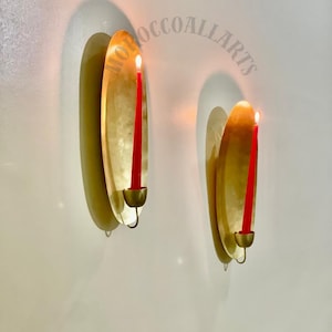 May include: Two wall-mounted candle holders with a gold finish. Each holder features a red candle, lit with a flame. The holders are mounted on a white wall, casting shadows. The text "MOROCCOALLARTS" is visible.