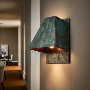 May include: A wall-mounted light fixture with a weathered, turquoise-colored shade and a warm, copper-toned interior. The fixture is rectangular and mounted on a matching metal plate. The light is on, casting a warm glow.