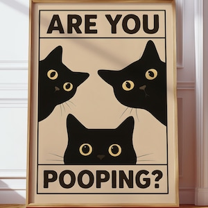 Può includere: Un poster incorniciato con tre facce di gatti neri con occhi gialli. Il testo "ARE YOU POOPING?" è visualizzato in lettere maiuscole nere e in grassetto. Il poster è in una cornice chiara, su uno sfondo neutro.