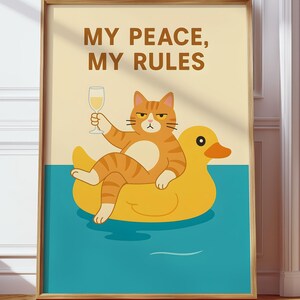 May include: A framed art print featuring a cartoon cat lounging on a yellow inflatable duck in blue water, holding a glass of wine. The print has the text "MY PEACE, MY RULES" in brown letters on a cream background.