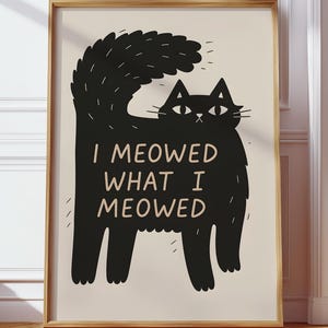 以下が含まれることがあります： 「I MEOWED WHAT I MEOWED」のテキストが入った黒猫のイラストが描かれた額入りのアートプリント。猫はふわふわの尻尾と厳しい表情をしています。アートワークはクリーム色の背景に設定されています。