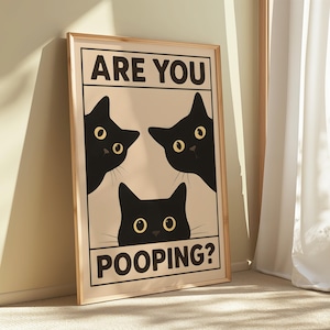 May include: A framed art print featuring three black cat silhouettes with yellow eyes on a beige background. The text "ARE YOU" is above the cats, and "POOPING?" is below. The frame is a light wood color.