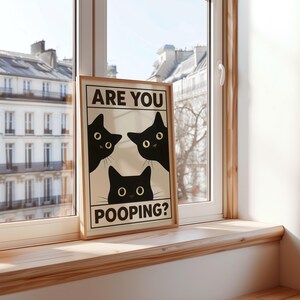 May include: A framed print featuring three black cat silhouettes with yellow eyes, peeking out from behind a white background. The text "ARE YOU POOPING?" is displayed in black, adding a humorous touch. The print is set against a window.