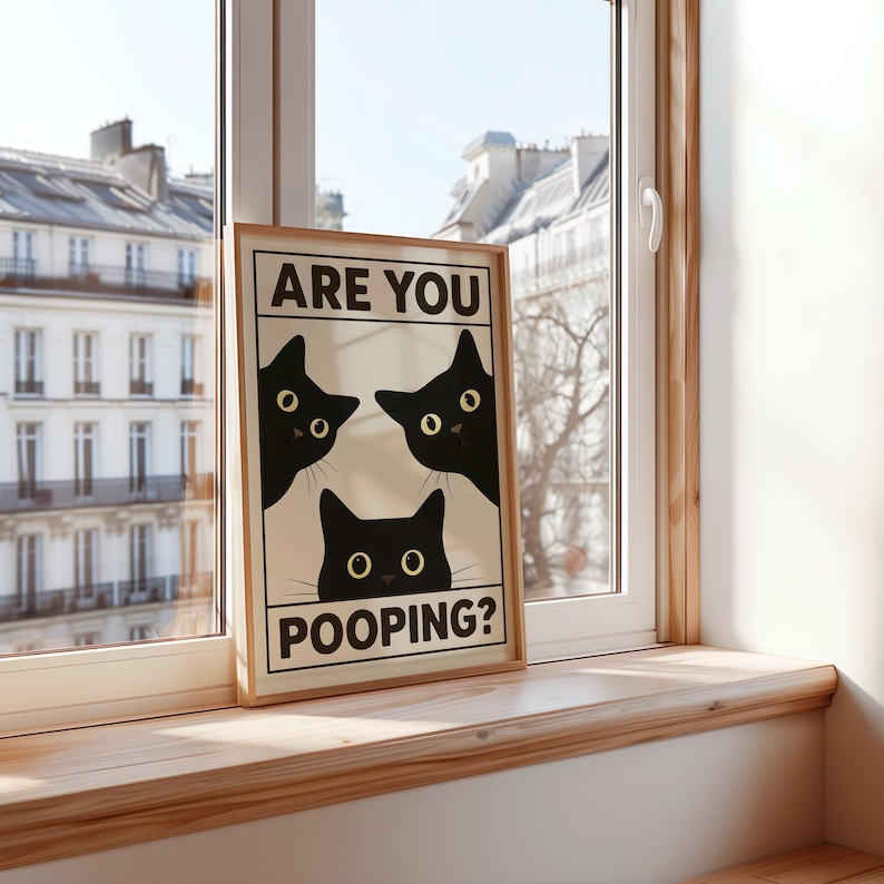 May include: A framed print featuring three black cat silhouettes with yellow eyes, peeking out from behind a white background. The text "ARE YOU POOPING?" is displayed in black, adding a humorous touch. The print is set against a window.