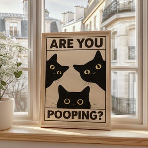 May include: A framed print featuring three black cat silhouettes with yellow eyes on a cream background. The text "ARE YOU POOPING?" is printed in bold, black capital letters. The artwork is displayed on a wooden windowsill.