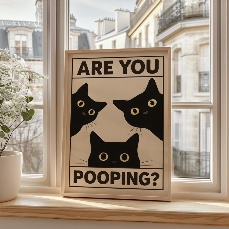 May include: A framed print featuring three black cat silhouettes with yellow eyes on a cream background. The text "ARE YOU POOPING?" is printed in bold, black capital letters. The artwork is displayed on a wooden windowsill.