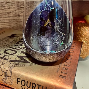 May include: A clear glass wine glass with a glittery silver rim. The inside of the glass is painted with a dark blue and black design with a lightning bolt pattern. The glass is sitting on top of a stack of books with the title "Fourth Wing" visible.