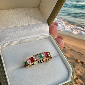 Könnte beinhalten: Ein goldfarbener Ring mit einer Reihe von bunten, rechteckigen Edelsteinen, darunter Rot, Rosa, Grün und Lila, wird in einer grünen Samtringbox präsentiert. Der Ring ist vor dem Hintergrund eines Strandes und Ozeans platziert.
