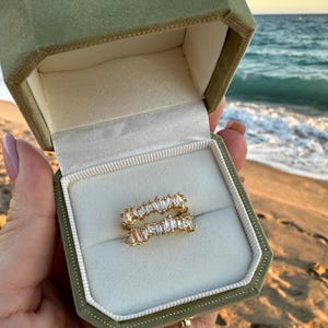 Peut inclure: Une bague en or ornée de pierres transparentes taillées en baguette est présentée dans un écrin en velours vert olive ouvert. L'écrin est tenu devant un fond de plage avec l'océan au loin.