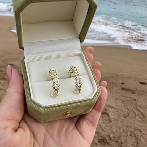 以下が含まれることがあります： 透明な宝石が付いた金のフープピアスが、開いた緑色のベルベットのジュエリーボックスに収められています。ピアスは、背景に海があるビーチの背景に配置されています。