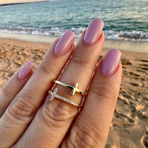 Puede incluir: Dos anillos dorados con diseños de cruces. Uno tiene una cruz con pequeñas piedras transparentes, el otro una cruz simple. Los anillos se llevan en un dedo con esmalte de uñas rosa, con una playa y el mar de fondo.