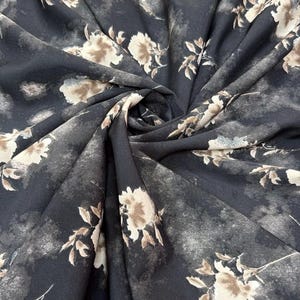 May include: A dark charcoal grey fabric featuring a repeating pattern of light beige and cream-coloured floral blossoms. The textile displays a soft, swirled drape with a matte finish, showcasing a watercolour-style print with muted, blended edges.