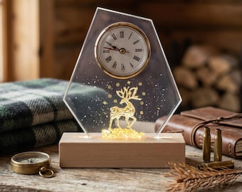 May include: A clear, pentagonal acrylic desk clock with a gold-rimmed face featuring Roman numerals. A gold-colored deer silhouette stands at the base inside the acrylic, surrounded by gold glitter. The clock rests on a rectangular wooden base.