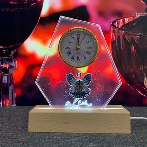 Può includere: Un orologio in vetro trasparente con un quadrante dorato e un design di fata con gioielli blu. L'orologio è su una base di legno e ha una luce bianca sotto.