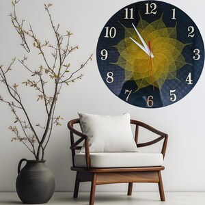May include: A round wall clock with a dark blue face and gold numerals. The clock features a yellow leaf design in the center. The clock is hanging on a white wall above a wooden chair with a white cushion and pillow.