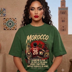 Peut inclure: Un t-shirt vert forêt à manches courtes avec une impression graphique d'un joueur de football en maillot rouge portant le numéro 26, un trophée doré et le texte "MOROCCO", "TOGETHER FOR BE PART OF THE DREAM WEAR YOUR PRIDE SUPPORT MOROCCO IN 2026".