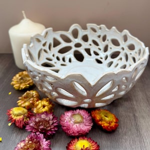 Puede incluir: Un cuenco de cerámica blanca con un diseño floral calado. Junto a él, una vela blanca y flores secas en tonos amarillos, rosas y rojos. El cuenco está sobre una superficie de madera.