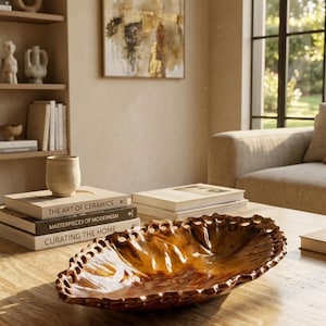 May include: A glossy, amber-colored ceramic bowl with a scalloped, perforated rim. The bowl has an elongated, organic shape and a smooth, reflective surface. It is positioned on a light-colored stone table next to a stack of three books.